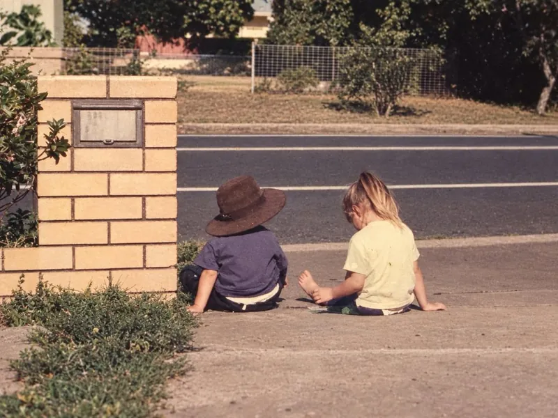 Emilee and Toby as little kids sitting on a footpath, Toby in a wide-brim hat